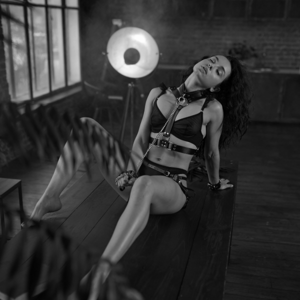 Schwarzweißaufnahme einer Person in künstlerischer Pose, mit Lederaccessoires und Lichteinfall durch ein großes Fenster. Die Szene vermittelt Stärke, Selbstbewusstsein und ästhetische Studioatmosphäre.