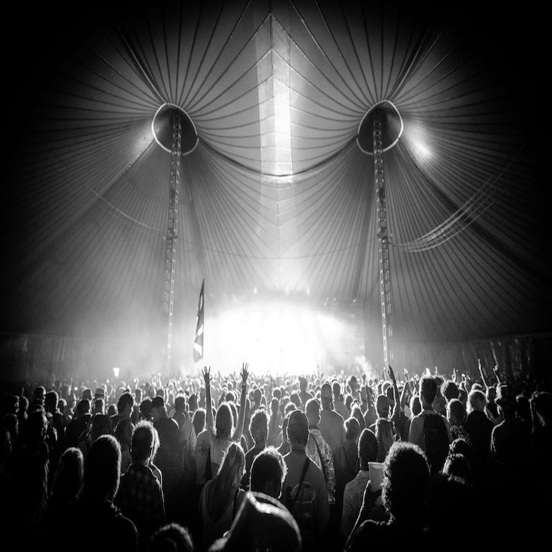 Schwarz-weiß-Aufnahme einer großen Menschenmenge bei einem Techno-Festival in einem Zirkuszelt mit spektakulärer Lichtshow – Arme in der Luft, euphorische Stimmung.