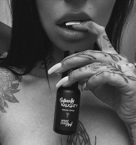 Mund und oberer Brustbereich einer tätowierten Frau mit gemachten Fingernägeln beißt sich sinnlich auf einen Finger und hält dabei ein Naturally Naughty Ingwerspray in der Hand. 